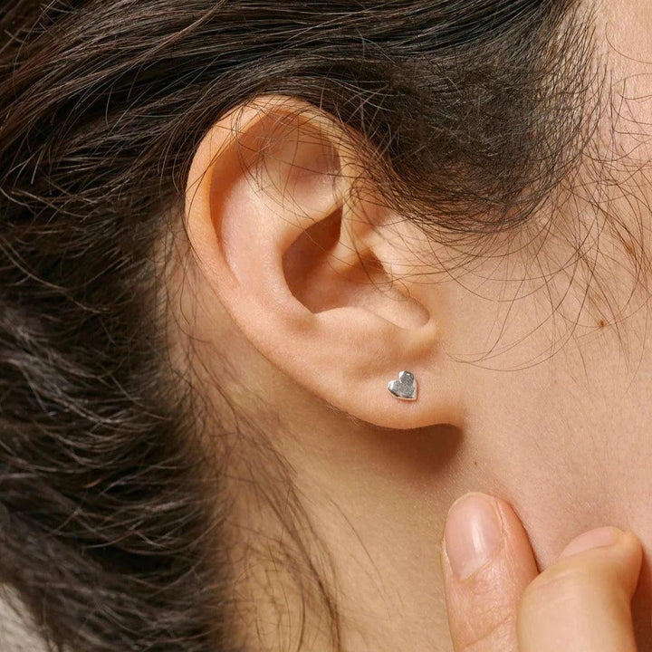 Enamel Heart Studs worden weergegeven als zilverkleurige hartjesoorbellen in het oor van het model, elegant en minimalistisch. ModelE373SM-E373SM-2