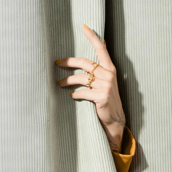 Vincent Orb Inferior vergulde ring, afgebeeld op de hand met gele nagellak voor een lichtgroen fluwelen gordijn. ModelVJ215-URG-001-2