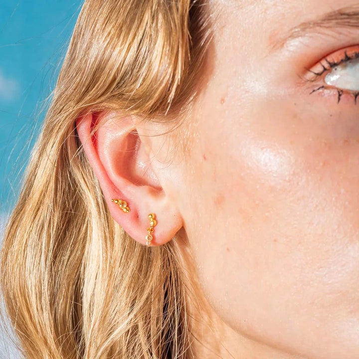 Vincent Pendulum Oorbellen Verguld weergegeven op het oor, kleine balletjes in goudlook onder heldere hemel. ModelVJ177-UEG-2