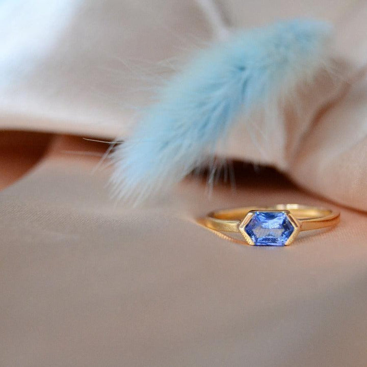anpe-atelier-op maat gemaakte-ring-met-blauwe saffier afgebeeld op beige stof, zeshoekige blauwe saffier in goud. Model27-blasafir-001-2