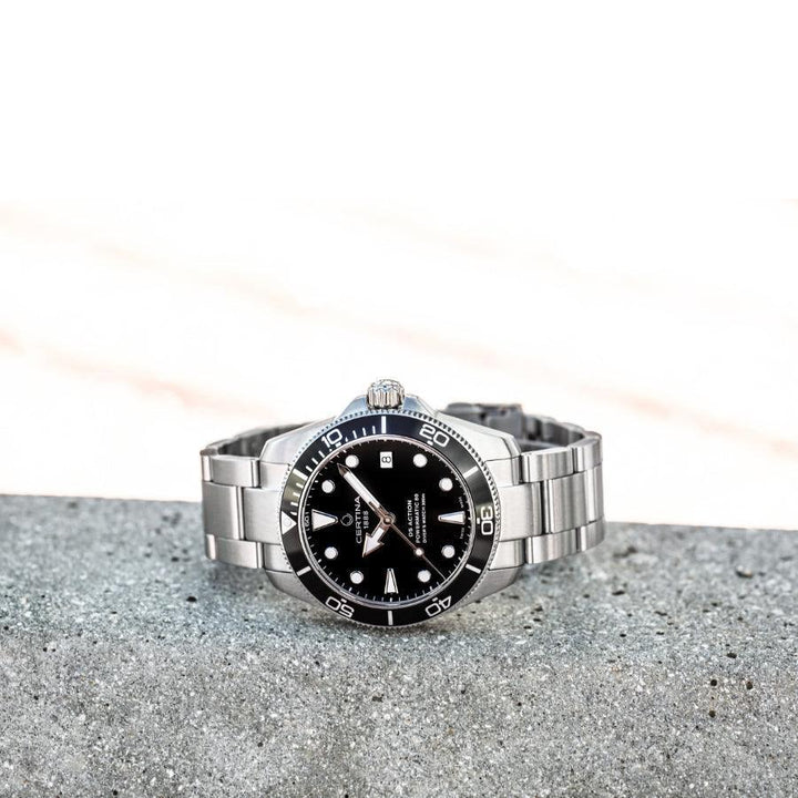 Certina DS Action Powermatic 80 herenhorloge met zwarte wijzerplaat en stalen ketting op grijs beton, model C0328071105100.